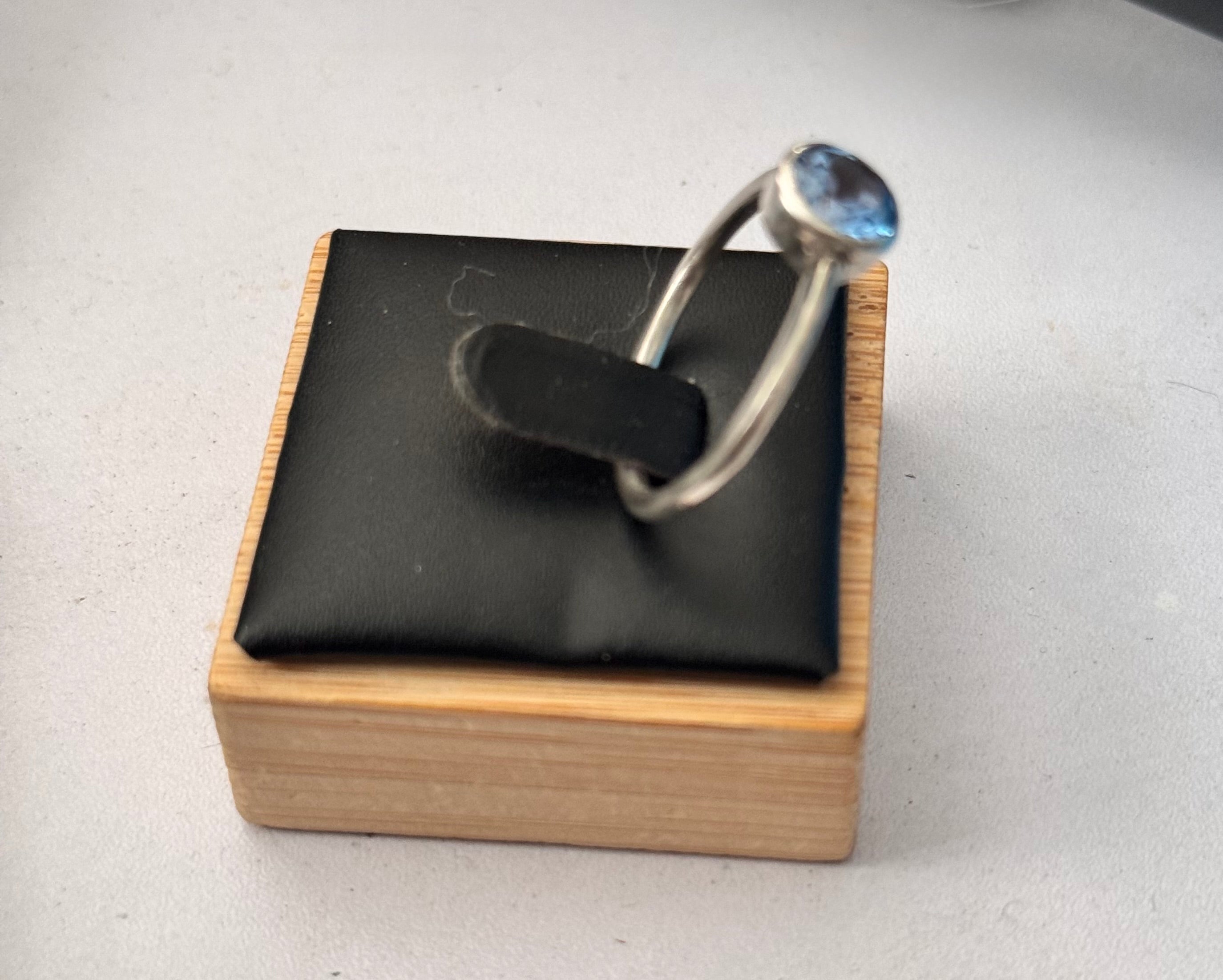 A silver ring with a round light blue gemstone displayed on a black cushioned wooden jewelry stand against a light gray background.