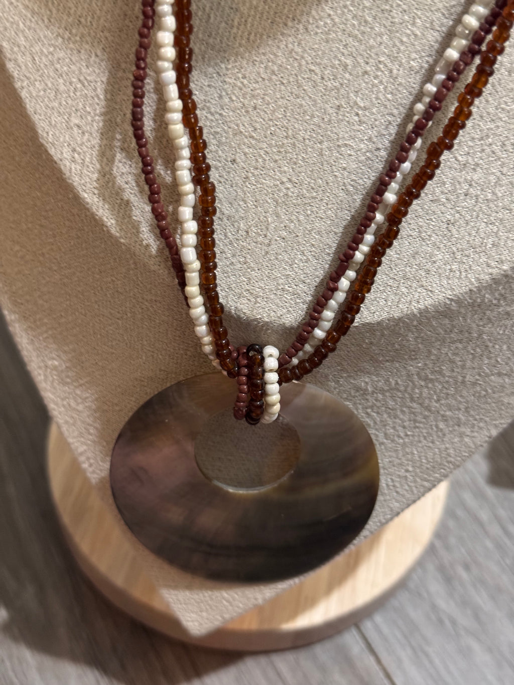 A beaded necklace with a large round shell pendant displayed on a beige jewelry bust. The necklace features multiple strands of small beads in dark brown, cream white, and reddish-brown, tied together at the top with a simple wrap.