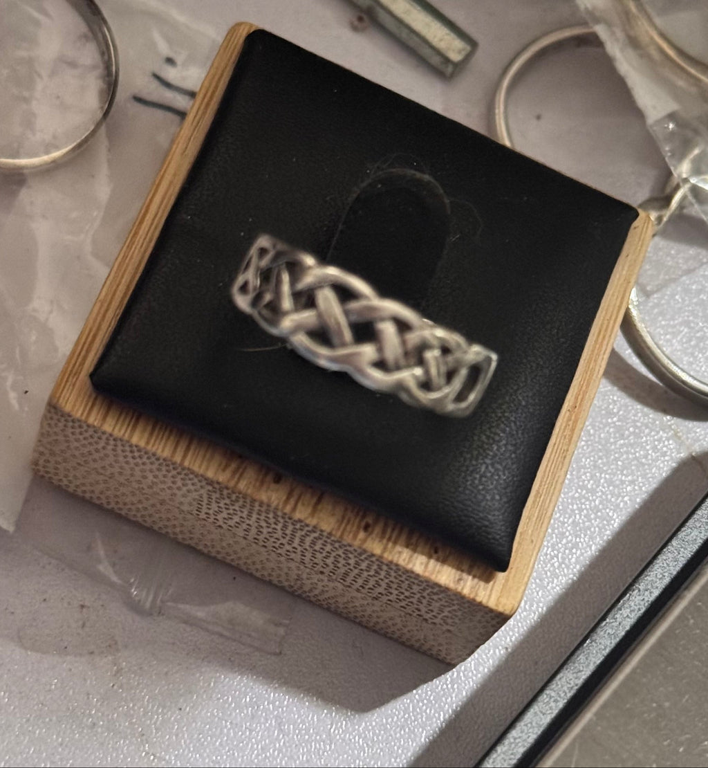 A close up shot of a sterling silver hallmarked, an interlaced Celtic knot ring displayed on a black cushioned wooden jewelry box, surrounded by other loose rings being prepared on a light gray surface.