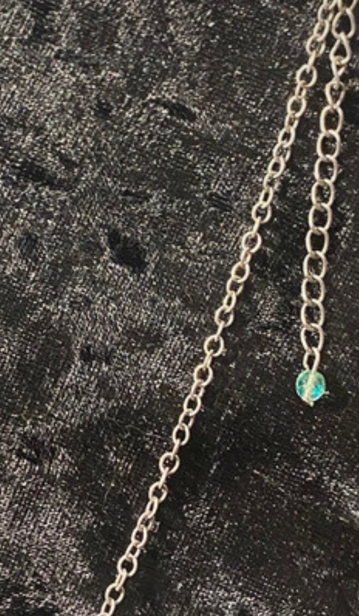A close-up of a silver chain necklace featuring a small, round, iridescent green bead charm, laid on a textured black fabric background. Thats the extender on the clasp