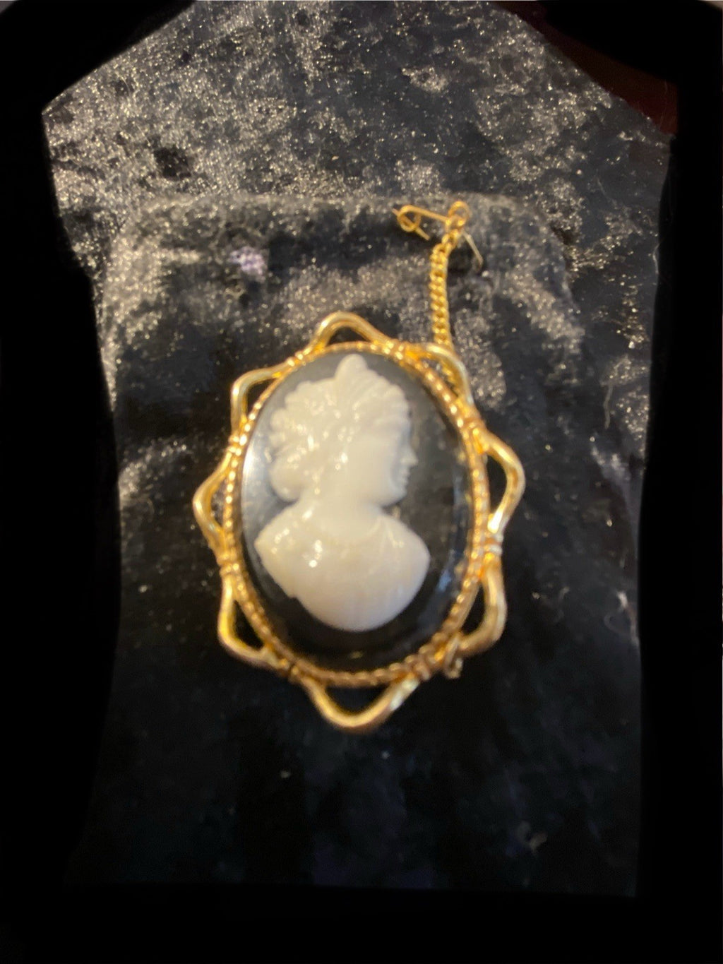 A vintage rolled gold cameo pendant featuring a white carved profile on a black oval background, displayed on a textured black surface.