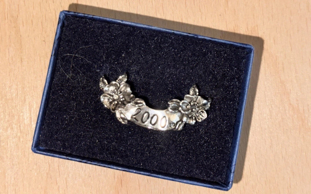 A silver-toned commemorative lapel pin with floral details and the year '2000' engraved on a black velvet jewelry box, placed on a light wood surface.
