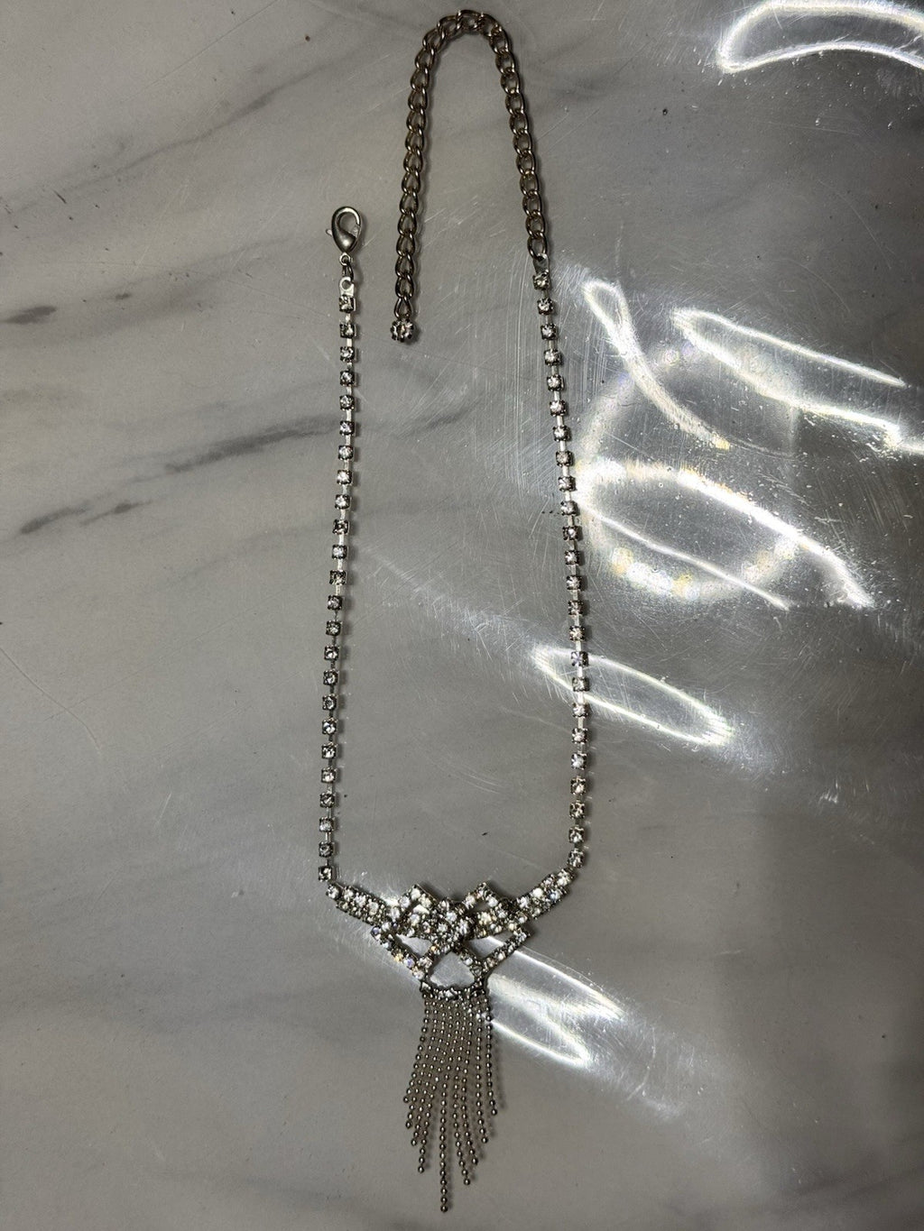 A silver-toned beaded necklace with a decorative crystal-embellished bow-like centerpiece and dangling chain fringe, laid flat on a light marble-like surface.