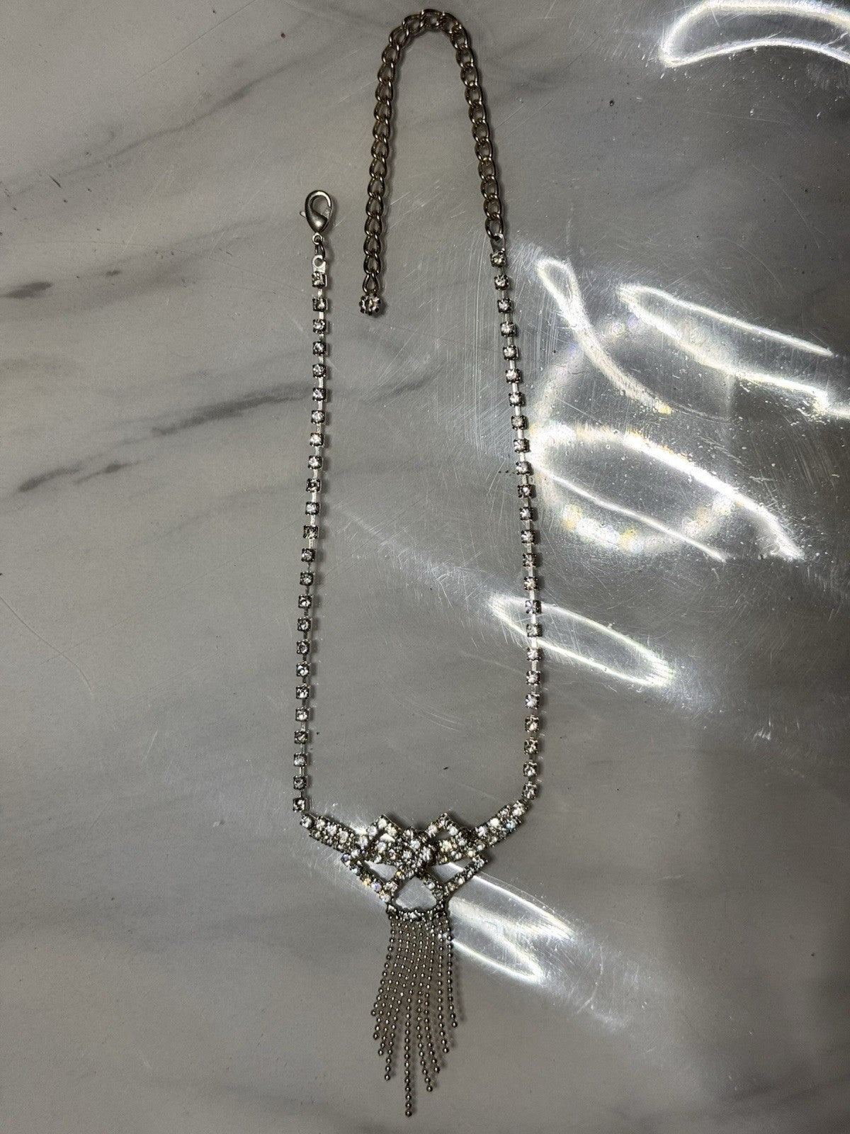 A silver-toned beaded necklace with a decorative crystal-embellished bow-like centerpiece and dangling chain fringe, laid flat on a light marble-like surface.
