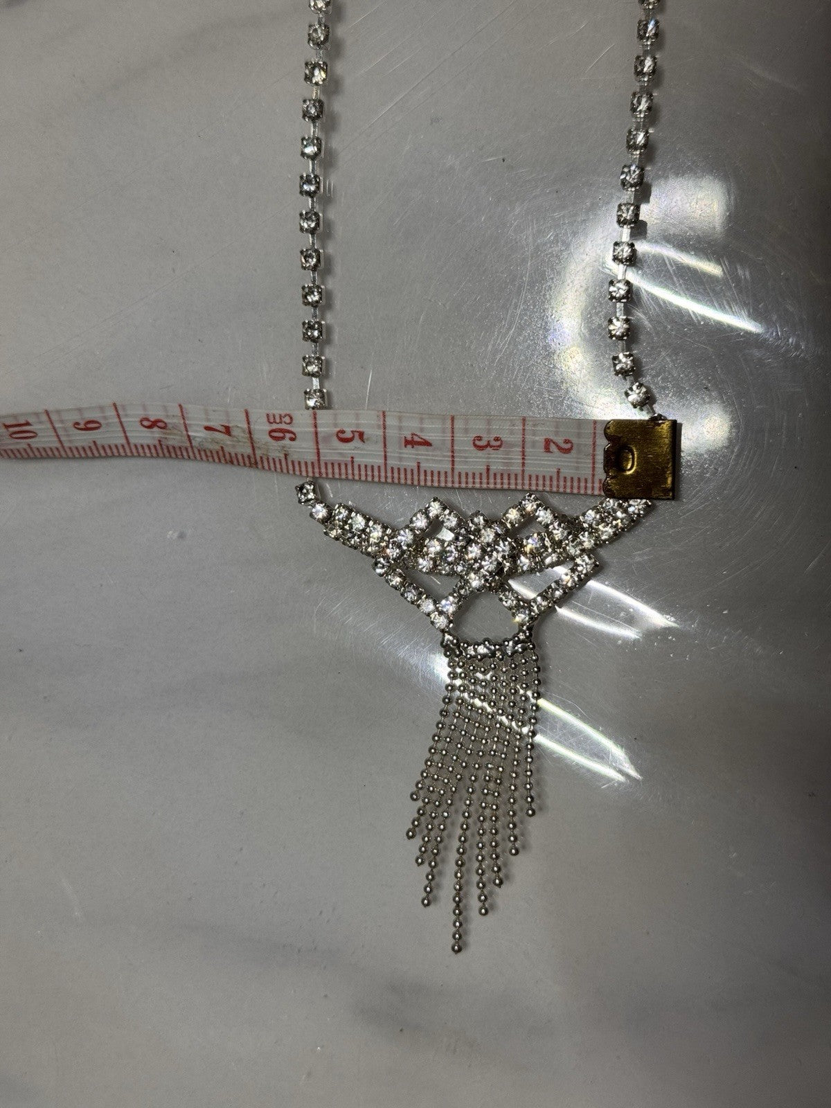 A silver-toned rhinestone necklace with a wing-like top and dangling chain fringe, shown on a metallic surface with a measuring tape across the upper middle for scale. T width of the bow part is 5 cms. Stunning statement bridal engagemnt necklace
