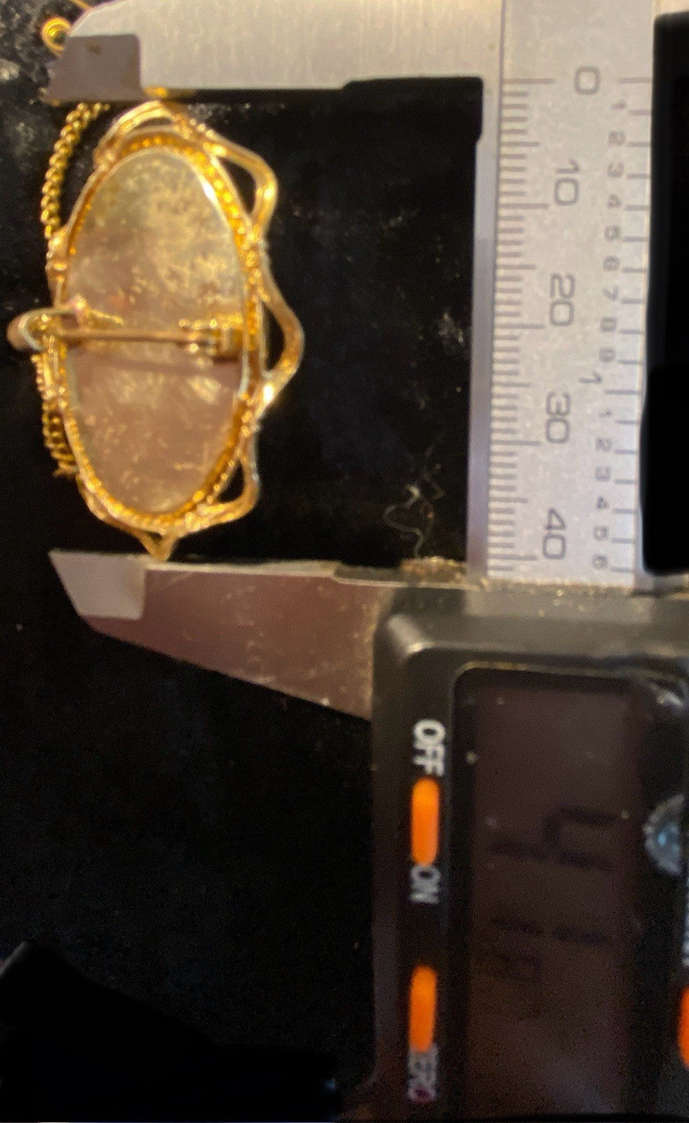 A  Rolled gold oval brooch or pin with a chain attachment, shown under a digital caliper against a black background. The caliper includes a metal ruler scale and a digital readout display recording the height of the cameo to be 41mm