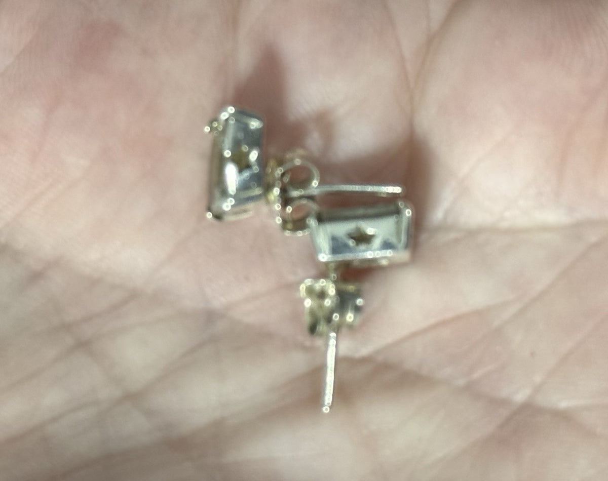 A pair of small silver-stud earrings with rectangular clear stones and a central star detail, resting on an open palm.