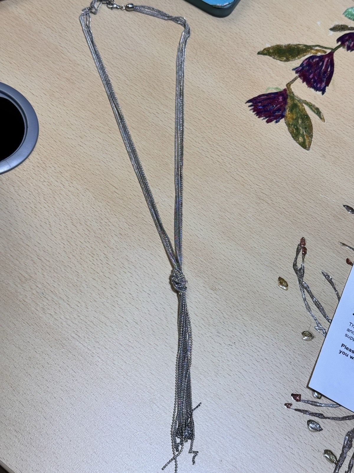 A long silver-toned chain necklace with a knotted center and tassel-like fringe, laid flat on a light wood surface with some decorative elements around it. The knot can be adjusted
