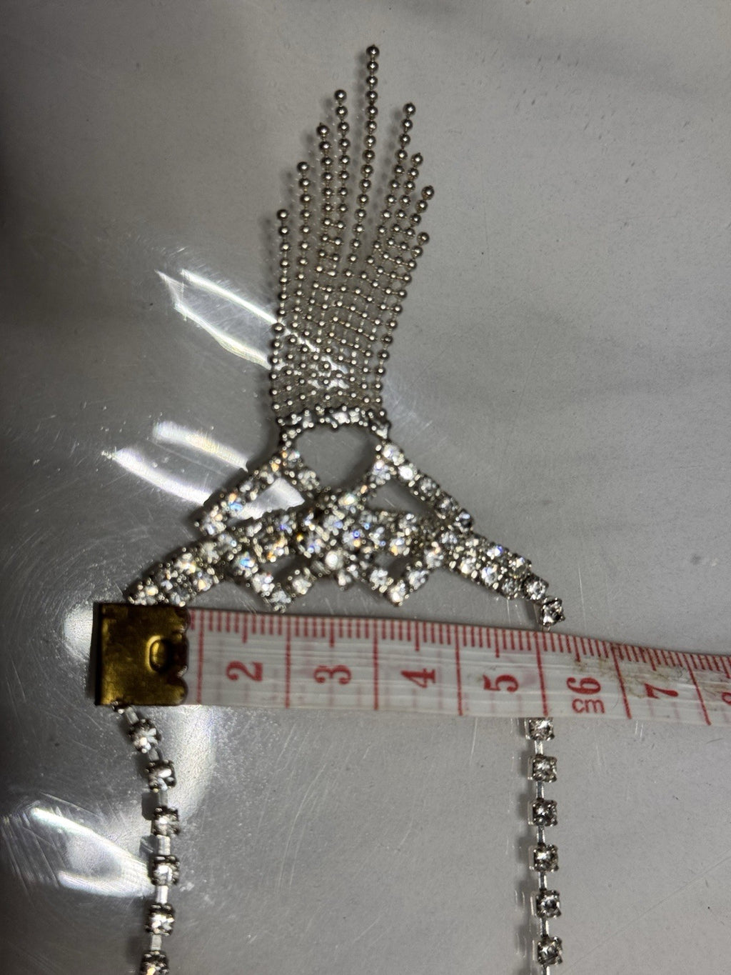 A silver rhinestone fringe necklace a close up of the fringe laid flat on a gray surface, with a measuring tape placed underneath the lower curved section to show scale. An beaty of the necklace