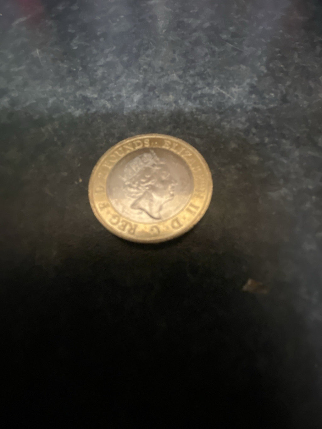 A small gold-colored 2 pounds British Coin with the queen, photographed on a dark textured surface.The other side is William Shakespear Sword in the crown.
Collectible coin