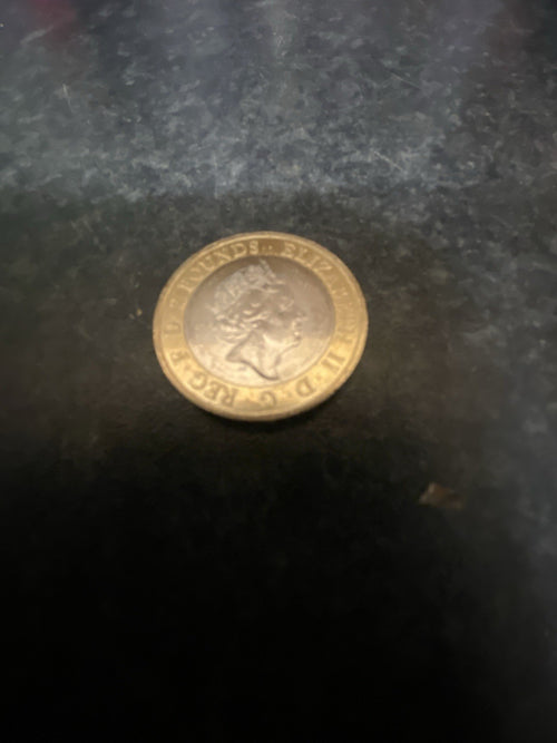 A small gold-colored 2 pounds British Coin with the queen, photographed on a dark textured surface.The other side is William Shakespear Sword in the crown.
Collectible coin