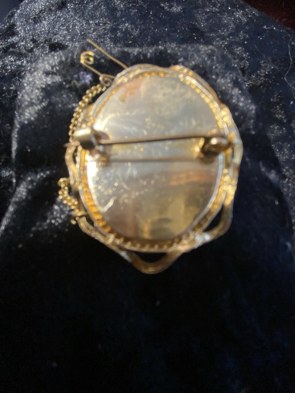An antique oval brooch or pin with a rolled - gold safety pin clasp and chain, featuring a textured, black and white-cameo center, displayed on a dark velvet surface.
