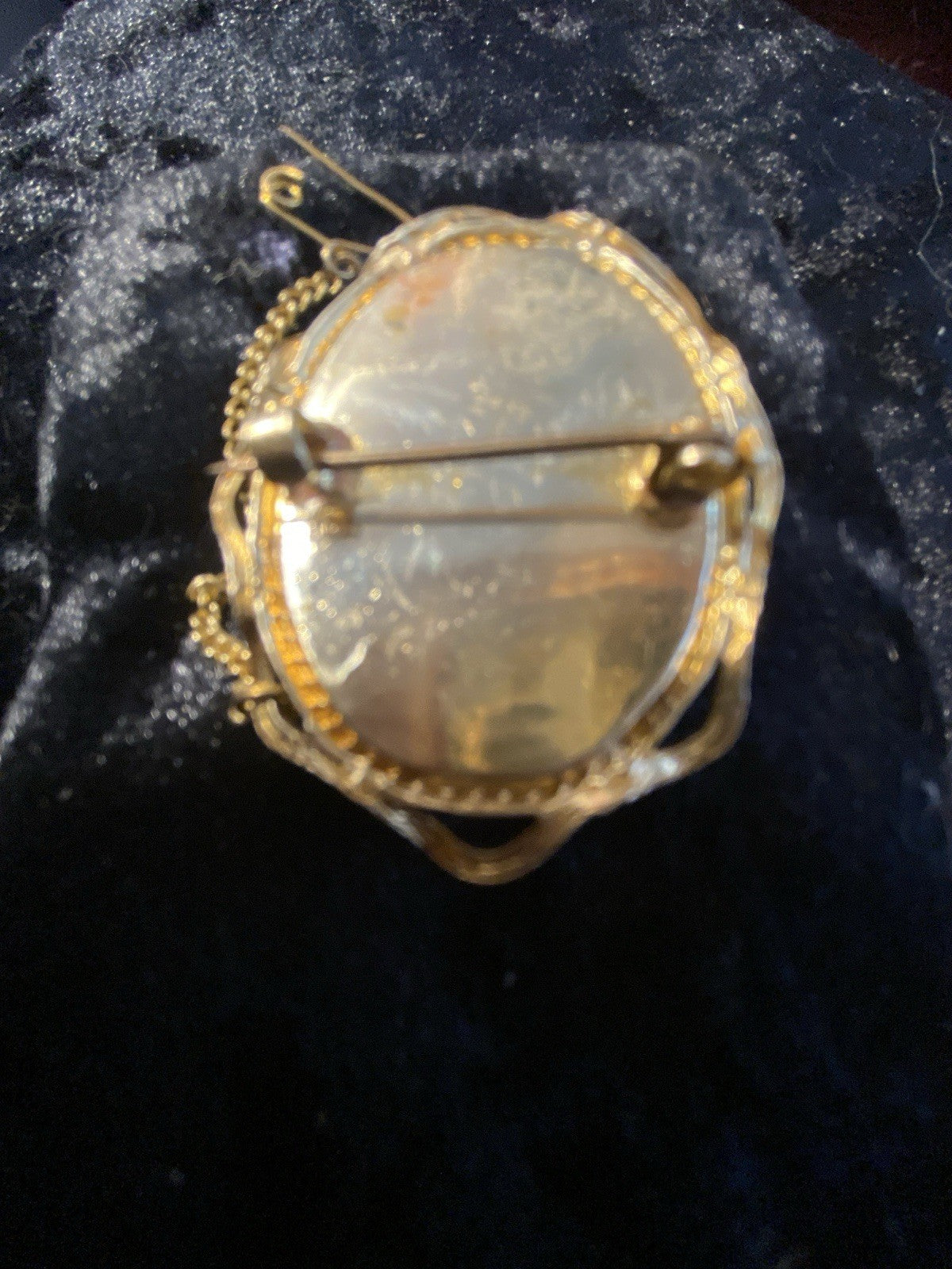 An antique oval brooch or pin with a rolled - gold safety pin clasp and chain, featuring a textured, black and white-cameo center, displayed on a dark velvet surface.