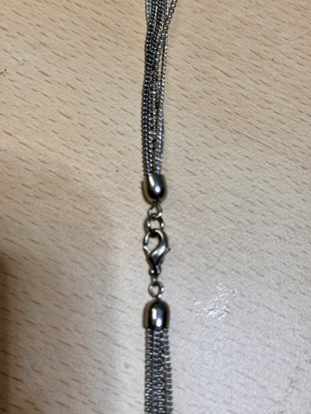 Close-up of a silver-toned chain accessory with a lobster clasp, bead details, and a tassel-like fringe on a light wood surface.