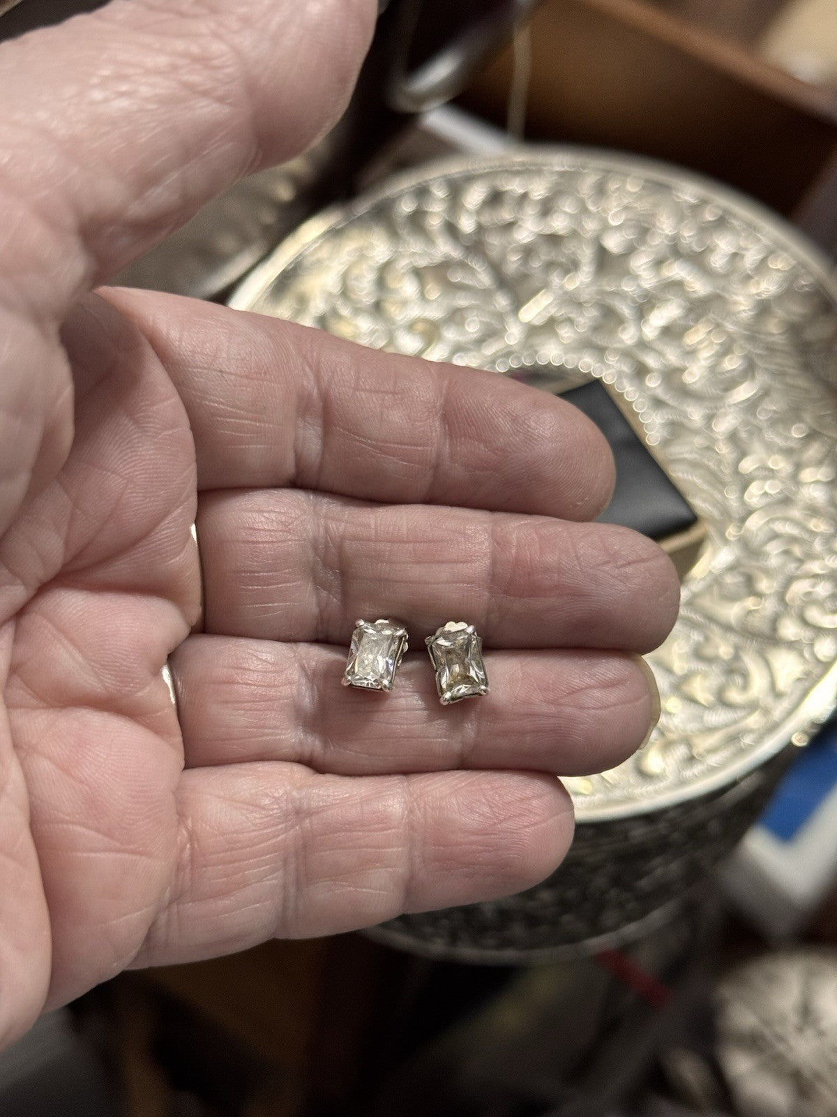 A close-up of a person's hand holding a pair of small silver stud earrings with clear rectangular-cut gemstones, with a decorative silver-toned container in the blurred background.