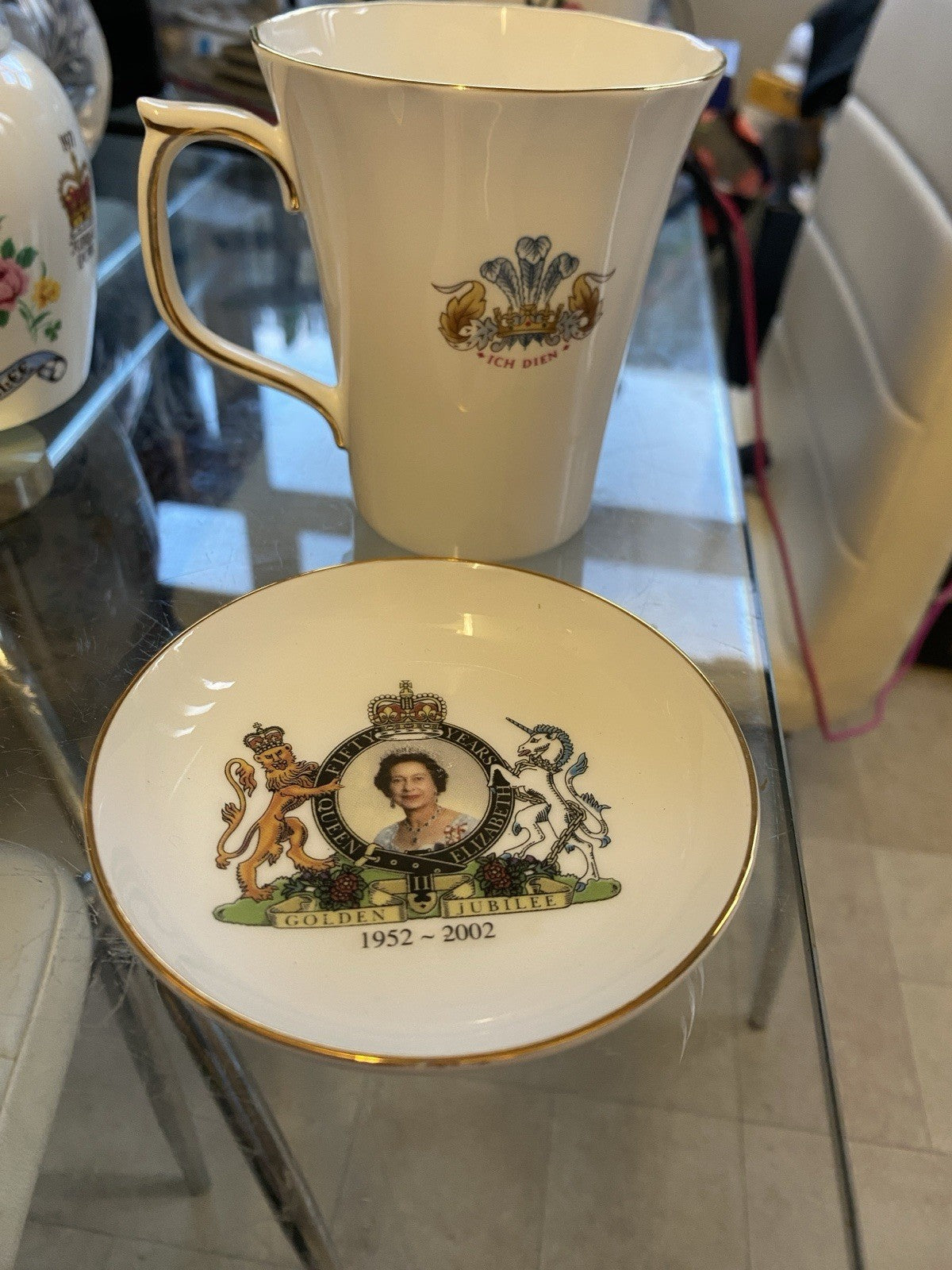 A white porcelain Princess Diana and saucer set with gold trim and a detailed Queen Elizabeth II Golden Jubilee on a glass table.