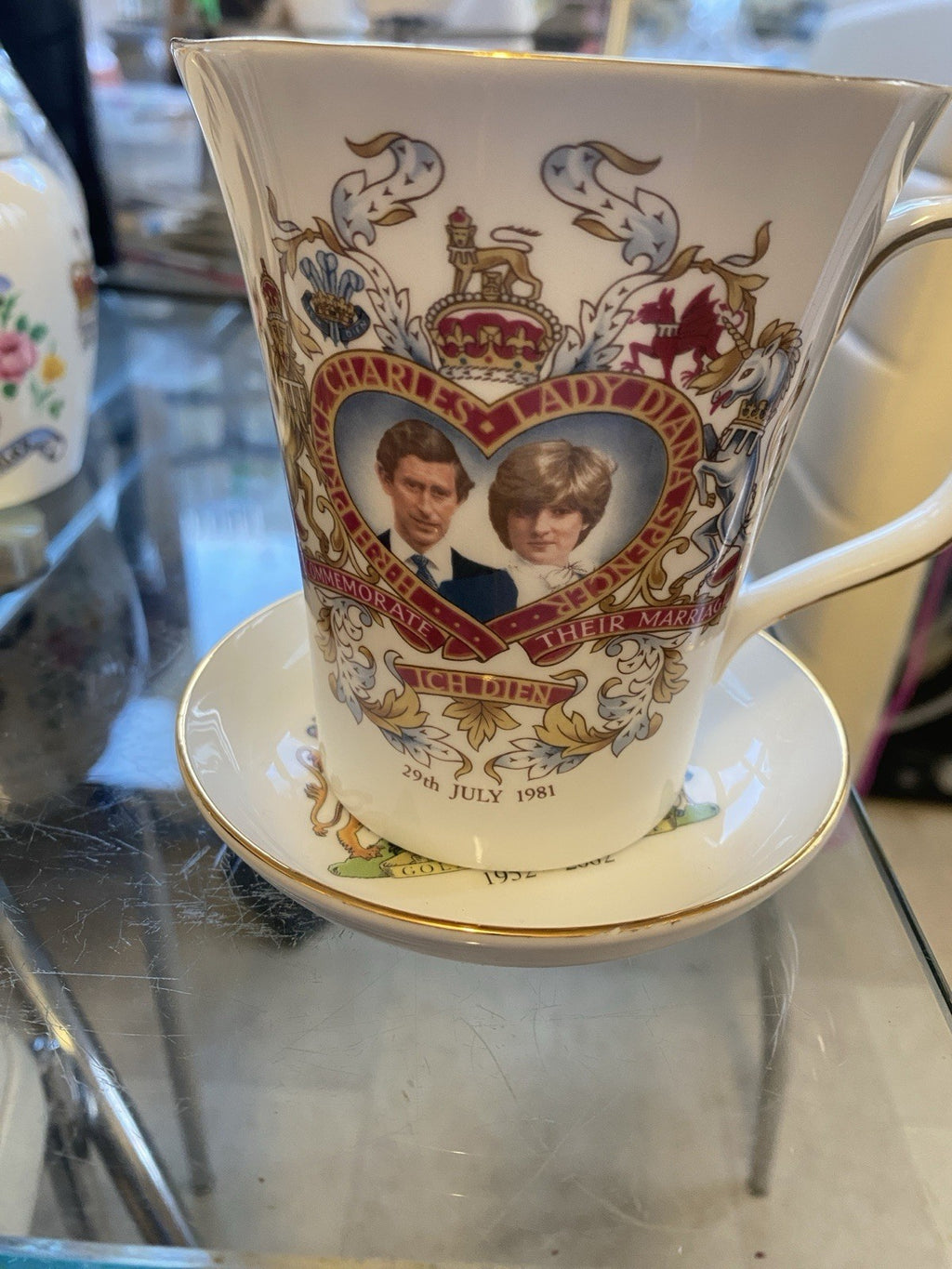 A white porcelain commemorative Mug and saucer featuring a printed portrait of Prince Charles and Princess Diana with ornate gold, red, and blue decorative detailing, displayed on a glass tabletop. This commemorates the Royal Wedding 1981
