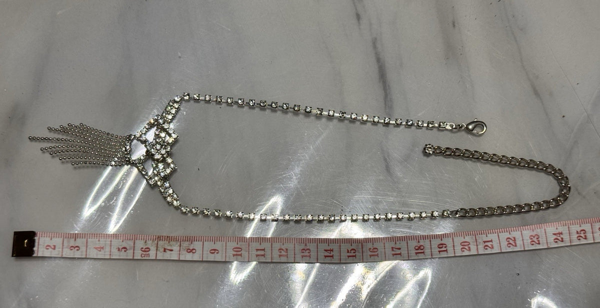 A silver-toned rhinestone necklace with a decorative tassel pendant, displayed on a light marble surface next to a measuring tape to show its length. the length closed is 24cms