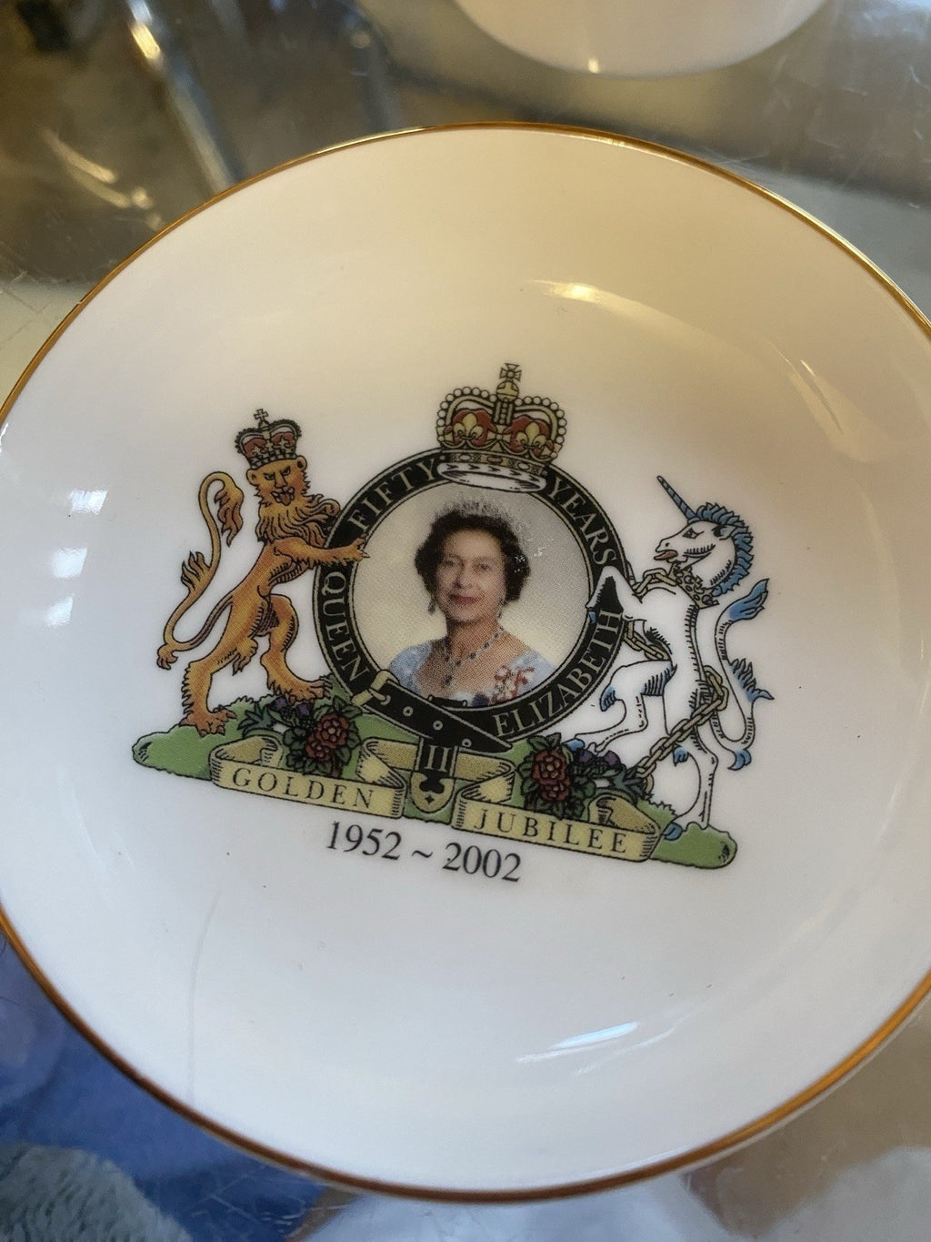 A white porcelain commemorative tea plate, saucer with a gold rim, featuring a portrait of Queen Elizabeth II inside a crown emblem, flanked by a lion and unicorn, with the text 'GOLDEN JUBILEE 1952 - 2002'.
