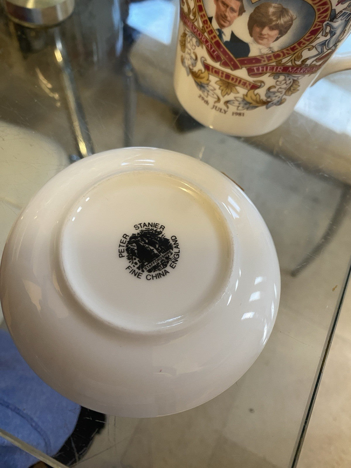 A white ceramic mug patially displayed behind the upturned saucer of the Queen Elizabeth 2nd's golden Jubilee, displaying a black printed circular logo that reads 'PETER STANIER FINE CHINA'. On a glass table