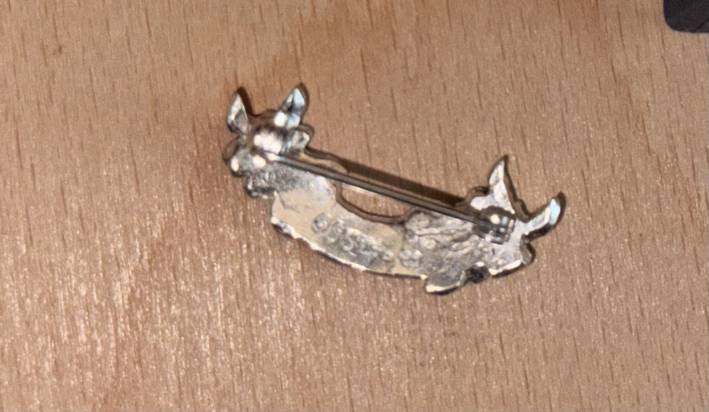 A small silver-tone metal pin or brooch showing the back side of the product and pin. Laid on a light wooden surface. English Rose
