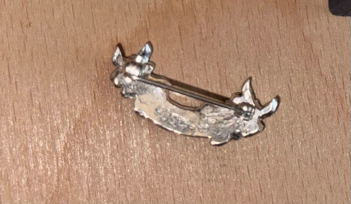 A small silver-tone metal pin or brooch showing the back side of the product and pin. Laid on a light wooden surface. English Rose
