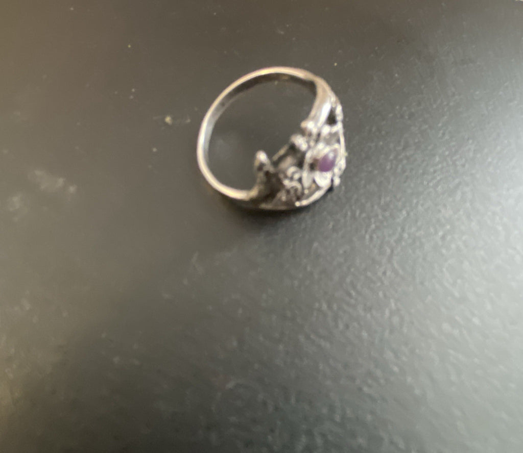 A silver-toned ring with a decorative filigree band and a small purple gemstone, resting on a dark gray textured surface.