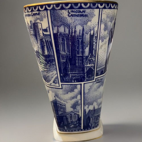 A blue and white illustrated ceramic Vase, Ringtons Tea Collectibles, Landmark Vase with blue architectural cityscape panels and gold trim, photographed on a light gray studio background with a cast shadow. This scene shows Lincoln Cathedral amongst scenes from Cornwall and other places