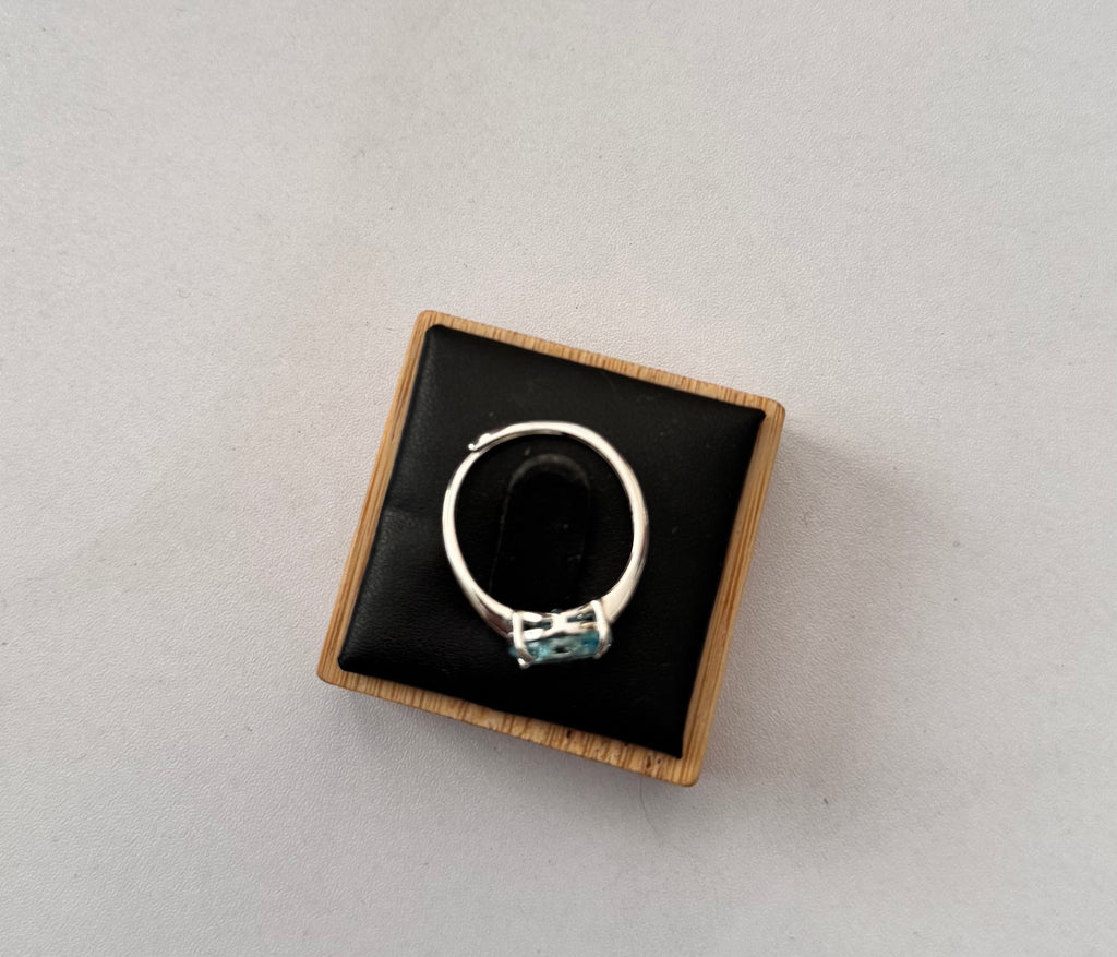 A sterling silver hallmarked ring with a small light blue gemstone set in a square wooden box with a black velvet interior, photographed on a light gray background.