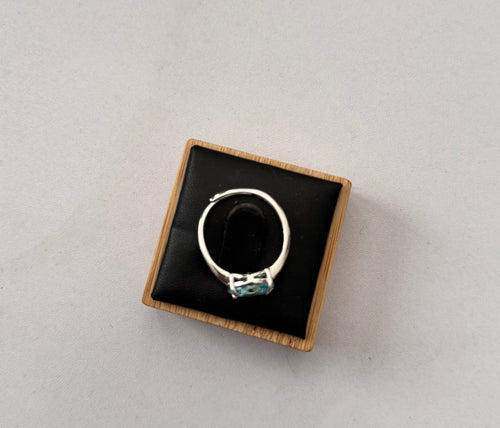 A sterling silver hallmarked ring with a small light blue gemstone set in a square wooden box with a black velvet interior, photographed on a light gray background.
