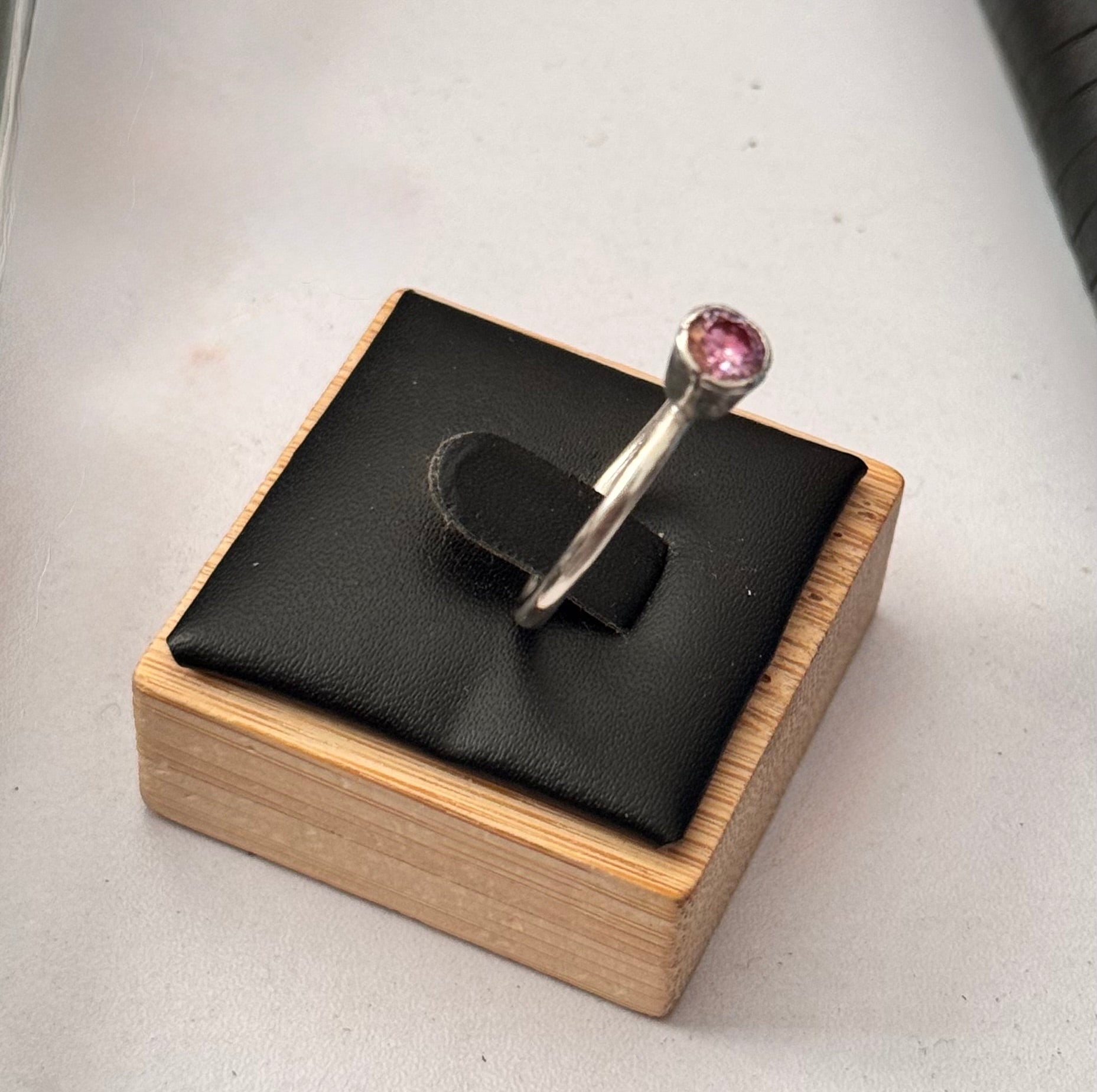 A silver ring with a round pink gemstone displayed on a black cushion set inside a wooden jewelry box, photographed on a light gray surface. Another view of the ring