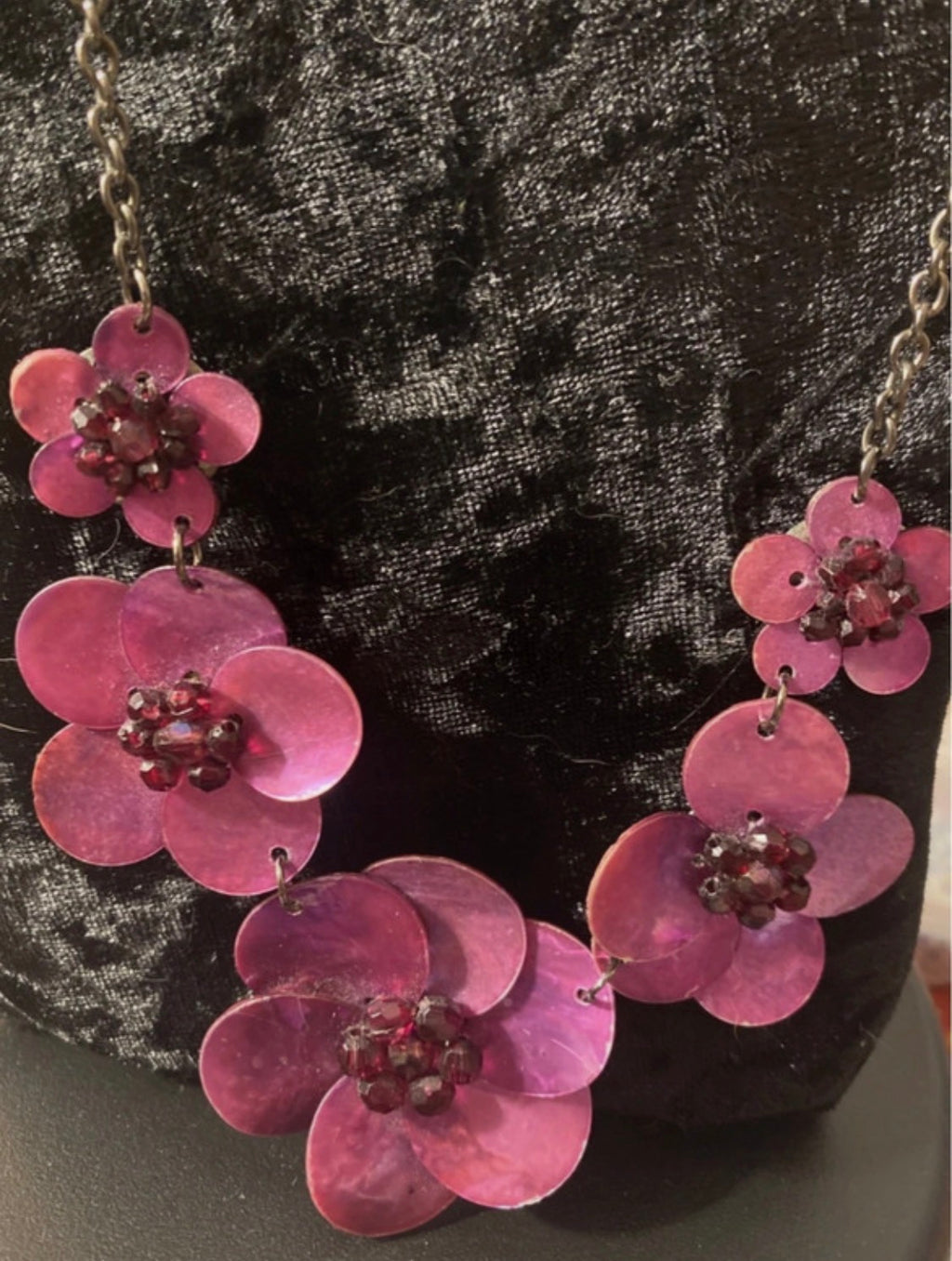 A floral statement necklace made of pink mother-of-pearl petals with dark red bead centers, displayed on a black velvet bust.