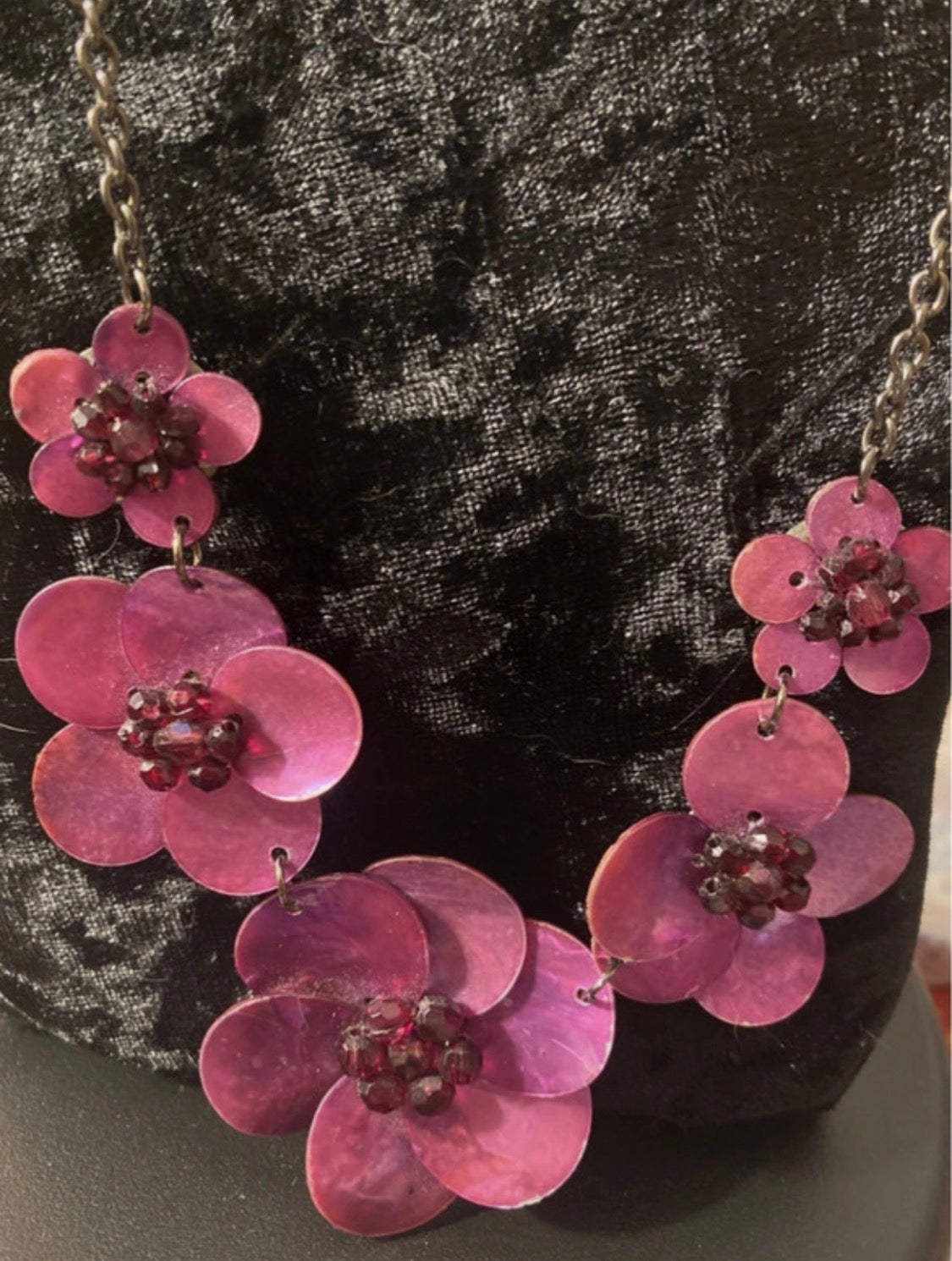 A floral statement necklace made of pink mother-of-pearl petals with dark red bead centers, displayed on a black velvet bust.