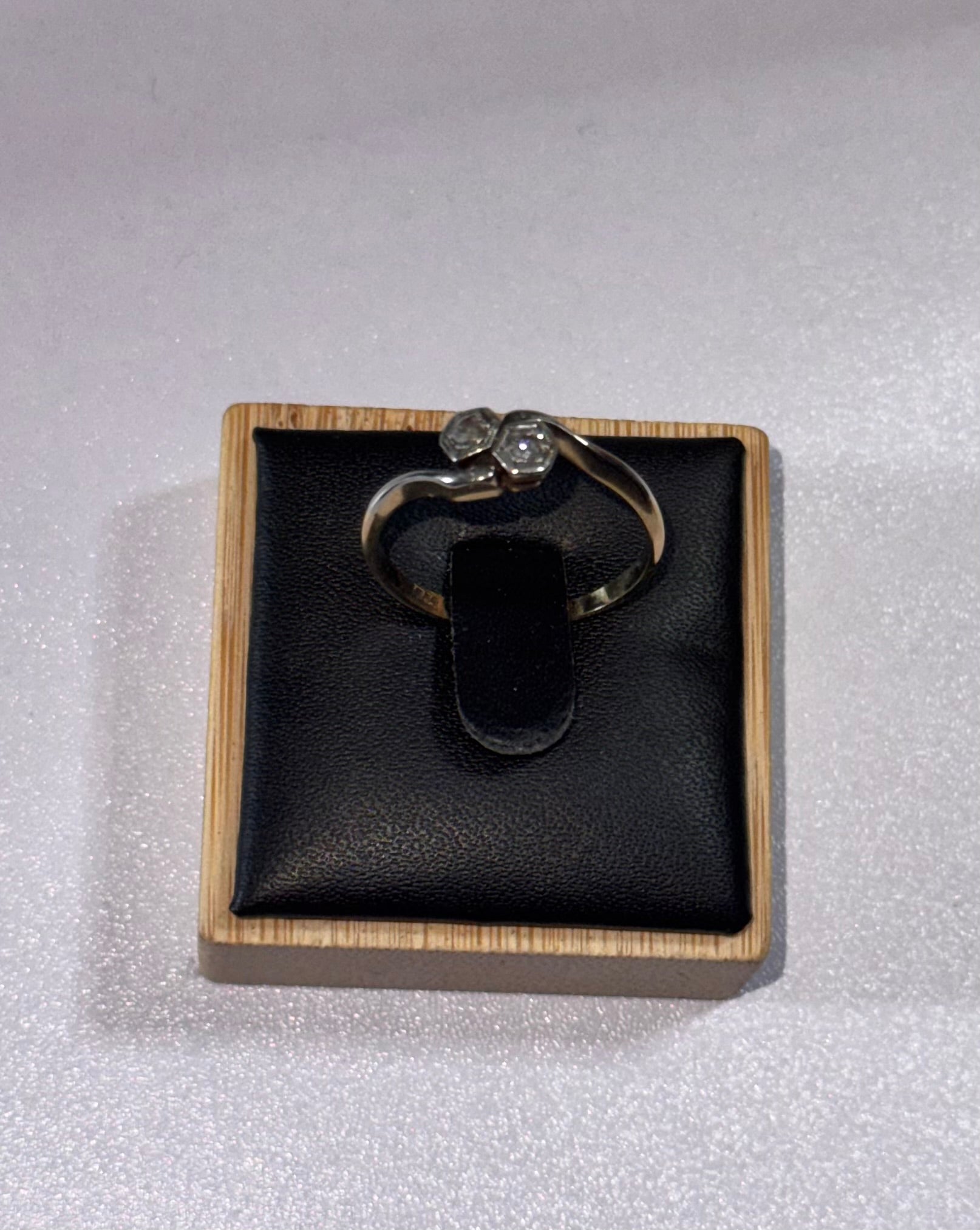 An 18 ct Gold ring with 2 diamonds in six sided ring settings, displayed on a black cushion inside a wooden box, set against a textured white background.