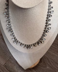 A silver-toned necklace with clear teardrop-shaped crystals displayed on a beige fabric jewelry bust against a wood floor background.