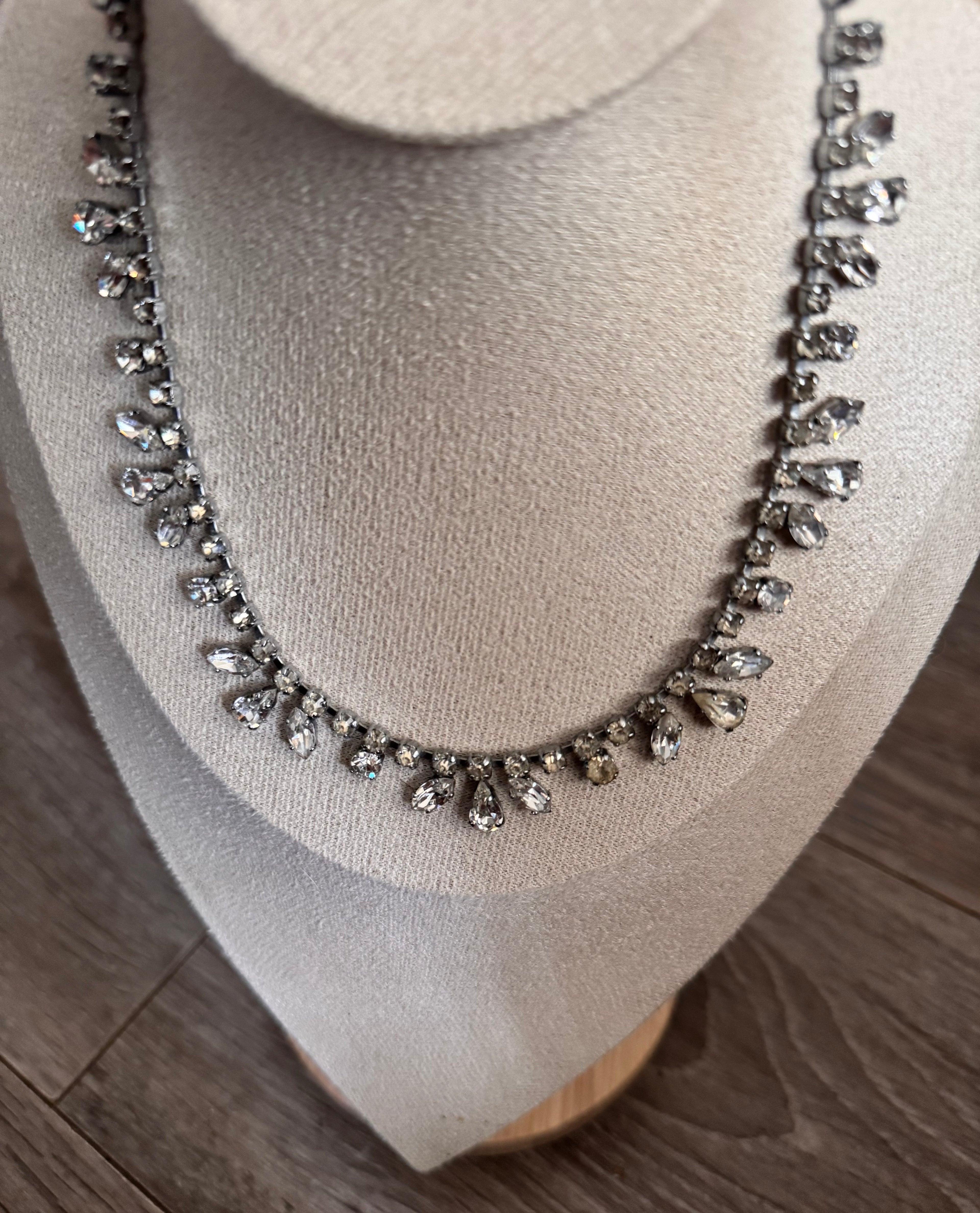 A silver-toned necklace with clear teardrop-shaped crystals displayed on a beige fabric jewelry bust against a wood floor background.