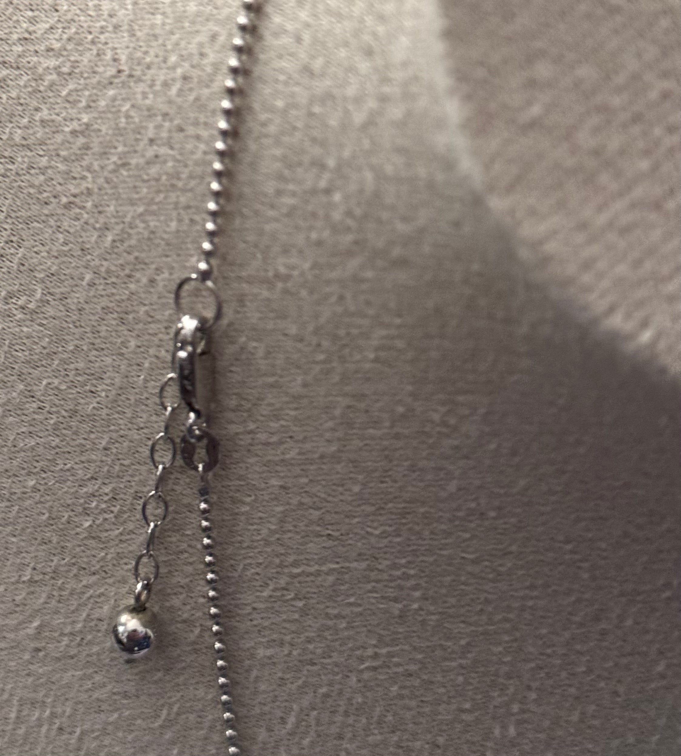 Close-up of a silver ball chain necklace with a lobster clasp and a small dangling charm, displayed on a textured beige surface.