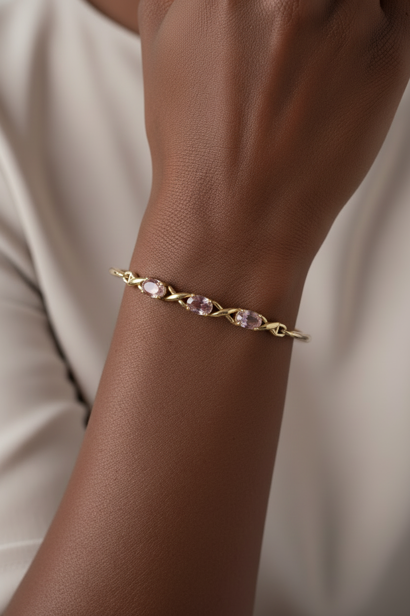 A sterling silver twist-link bracelet with three oval light pink/purple toned gemstones, shown worn on a person's wrist against a soft cream-colored top. Another image to display the bangle worn. It is a silver bracelet. Hallmarked and worn by a person of darker skin tone. Produced by Ai.