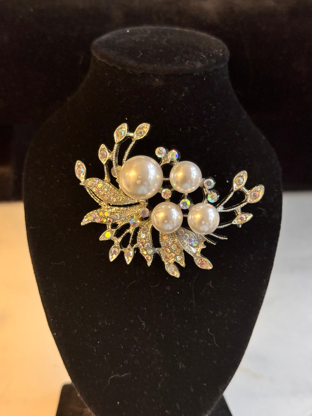 A floral brooch featuring four large white faux pearls and iridescent rhinestones on a silver-tone setting, displayed on a black velvet jewelry bust.