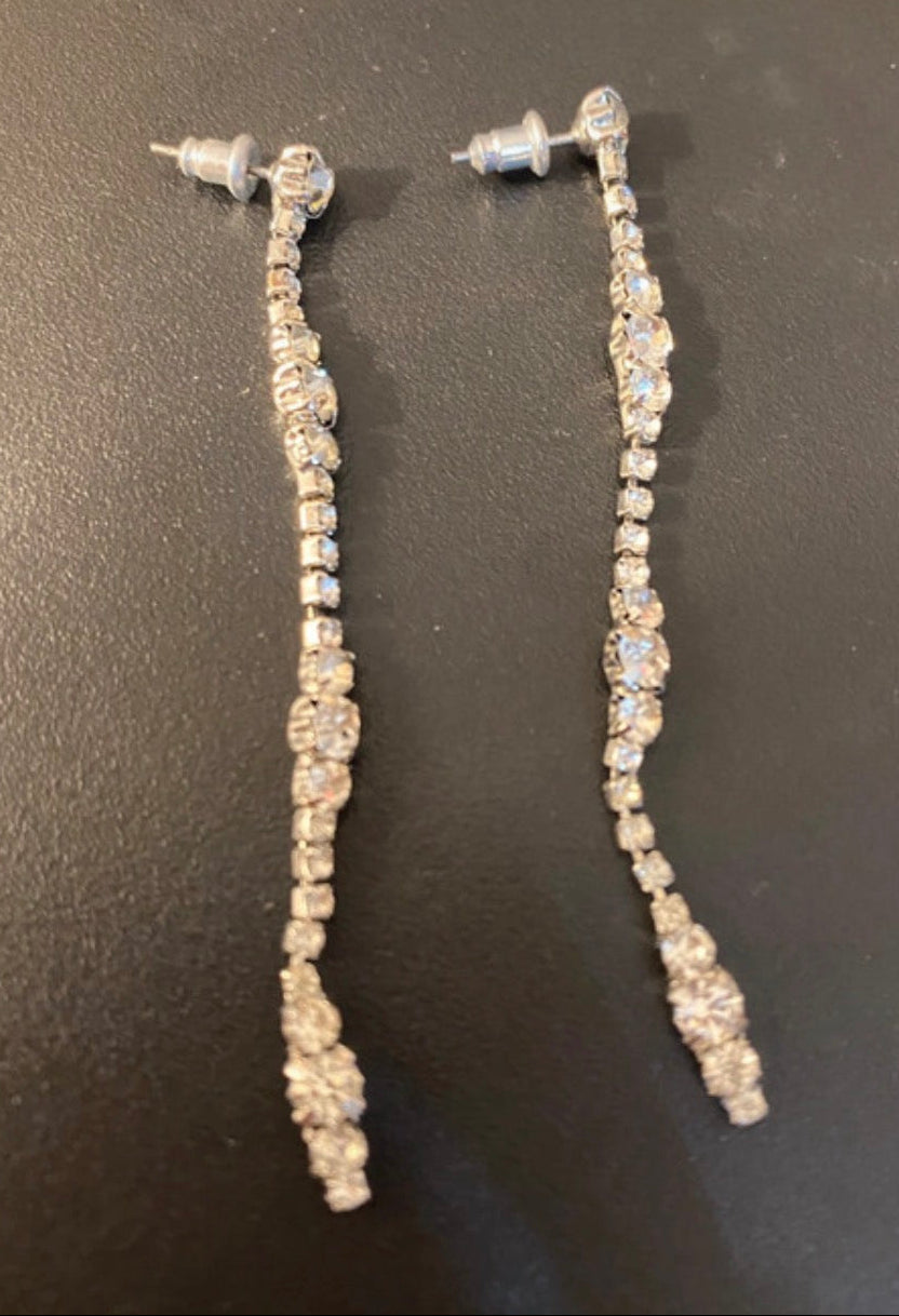 A pair of silver-toned dangle earrings with clear rhinestones, displayed on a dark gray textured surface.