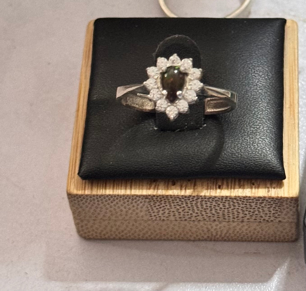 A sterling silver hallmarked ring with a dark oval center stone possibly moss agate as glows red in some lights and green in others is a striking  halo ring of small clear stones, possibly czs, displayed on a black cushion atop a light wood box.