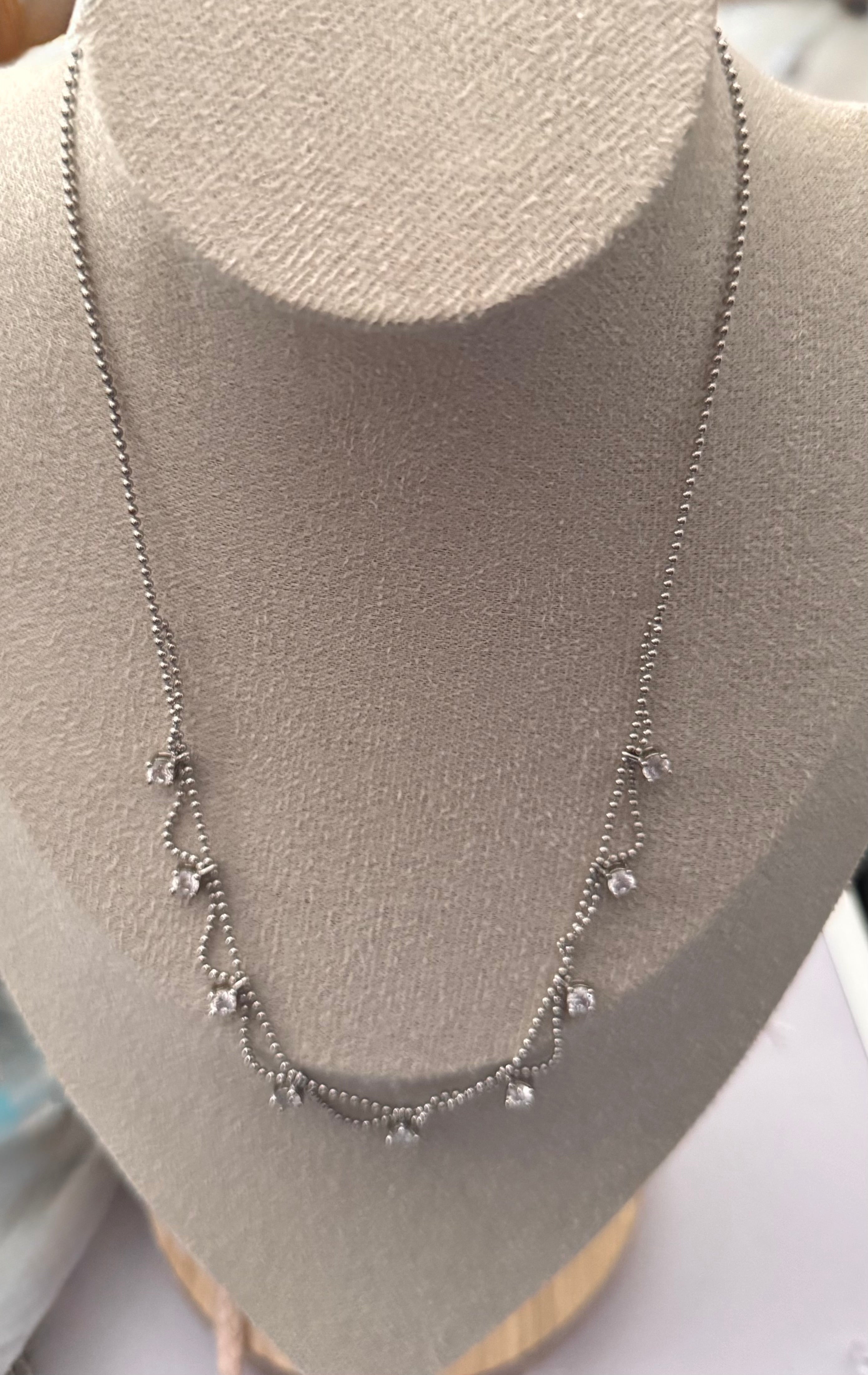 A delicate silver-toned necklace with small clear stones, displayed on a beige fabric jewelry bust.