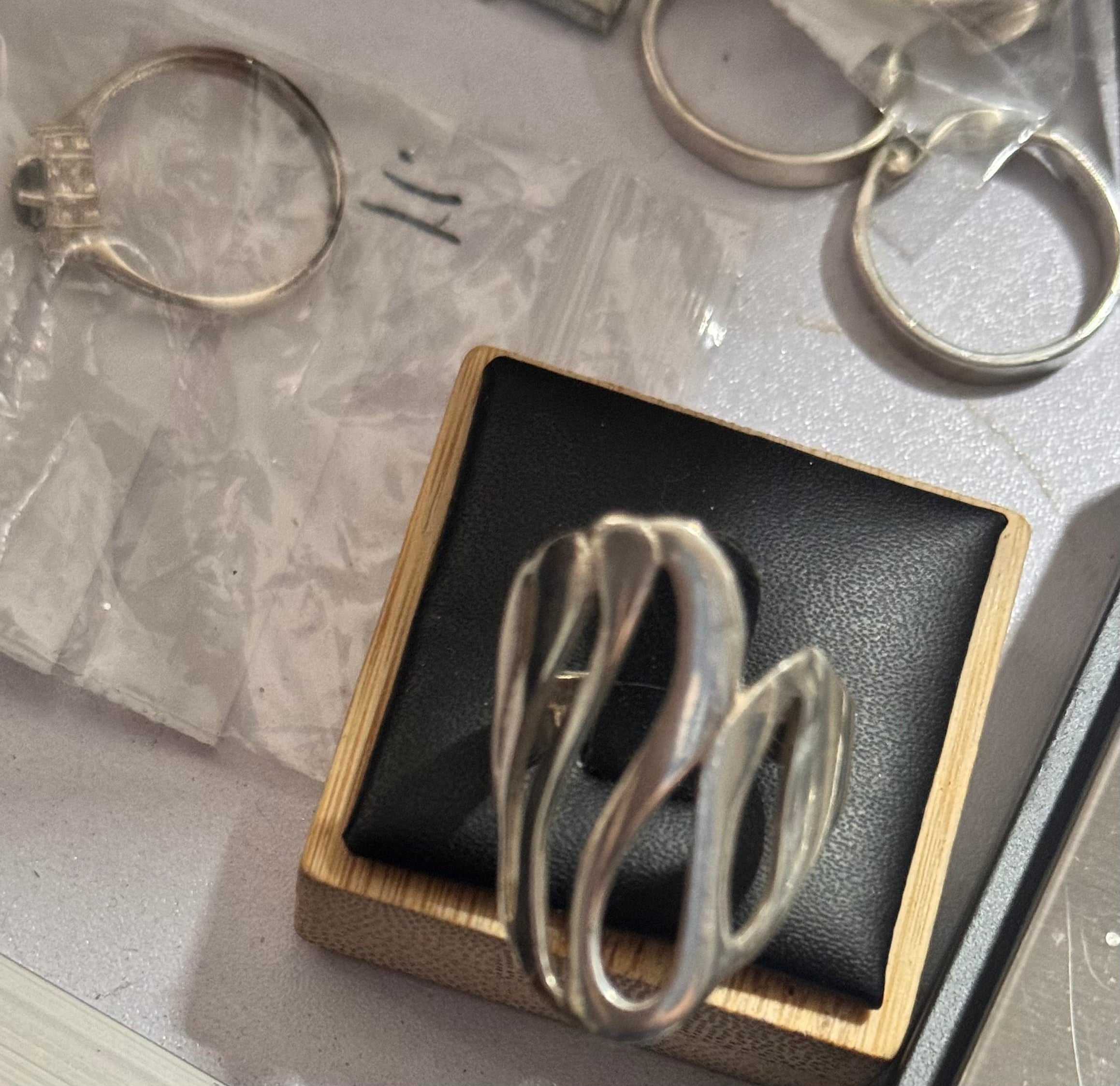 A close up of a sterling Silver wavy ring displayed on a black cushion inside a light wood box, with other rings and plastic bags in the background on a tabletop.