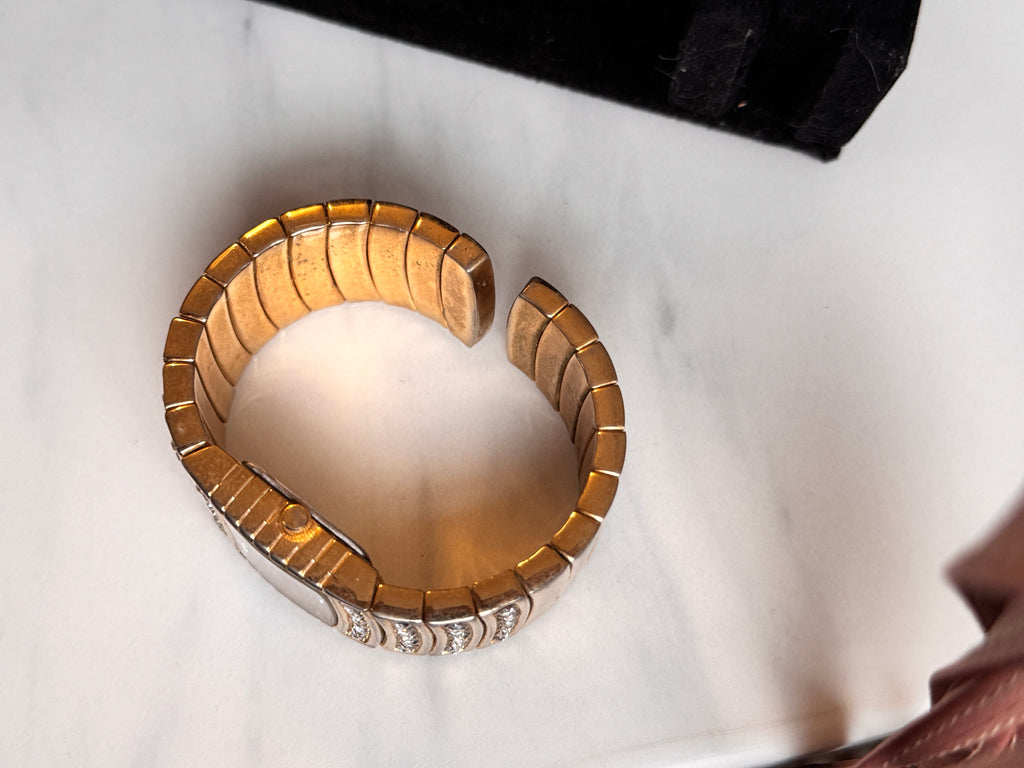A gold-toned segmented cuff bracelet watch set with small clear stones, photographed on a white marble-like surface. The watch expands slightly to place on the Wrist