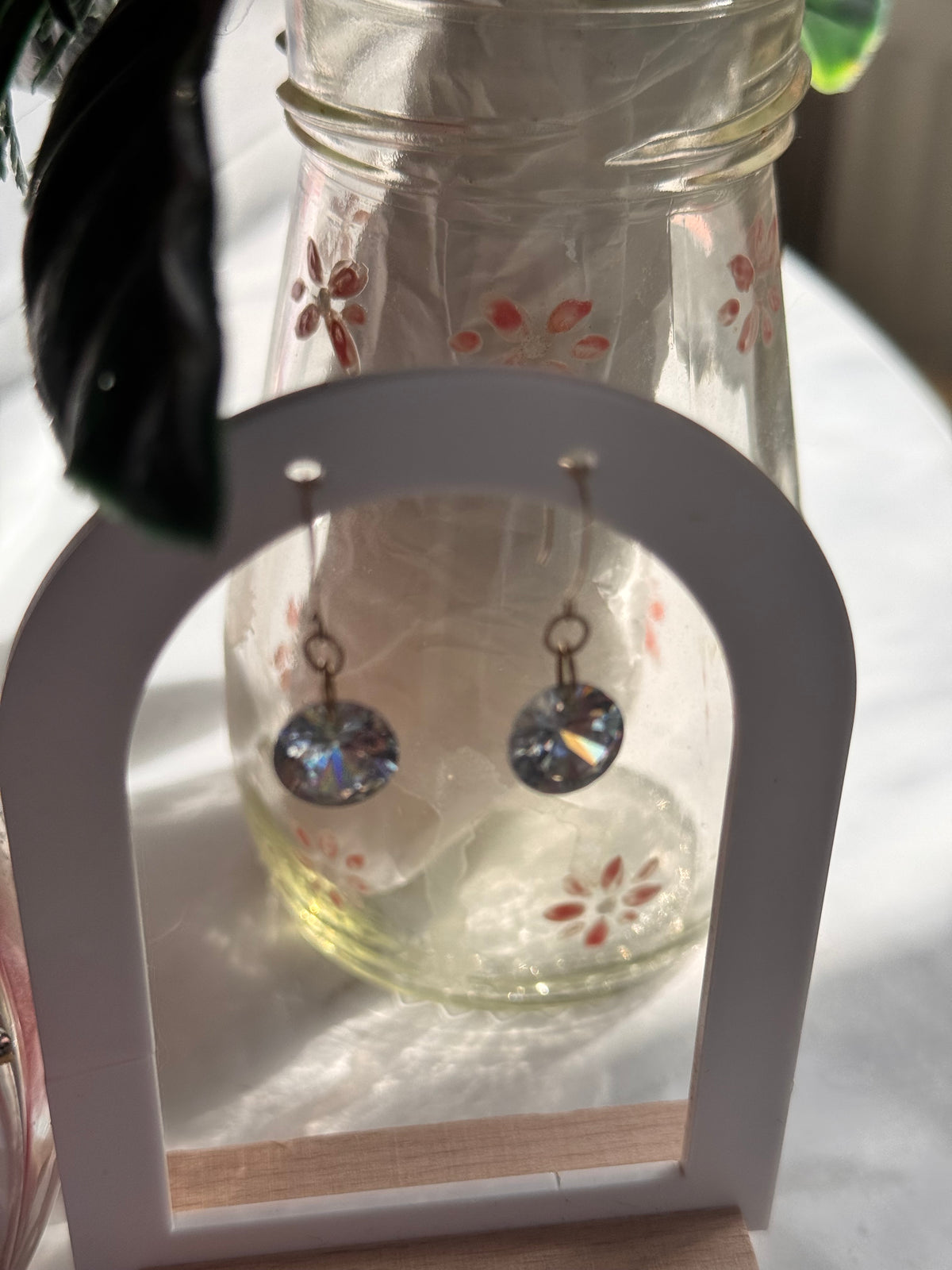 A pair of dangling earrings with round iridescent clear rainbow type faceted stones, displayed on a white arch stand in front of a clear glass bottle with red floral decals.