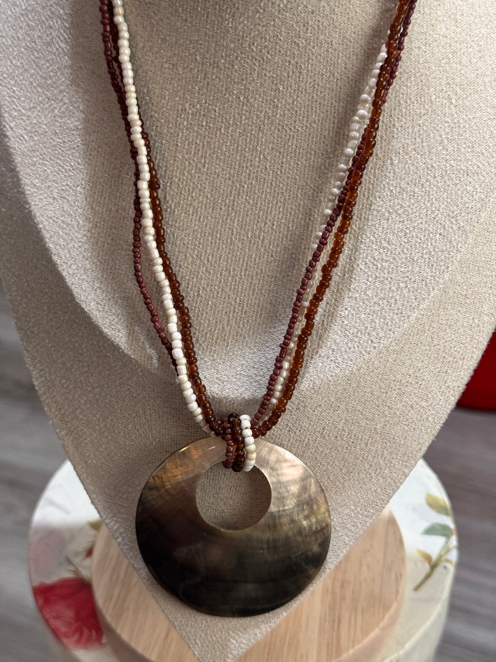 A bohemian-style necklace featuring a large circular shell pendant in a natural brown and black gradient, suspended from two beaded strands in dark brown and cream white, displayed on a beige fabric jewelry bust.