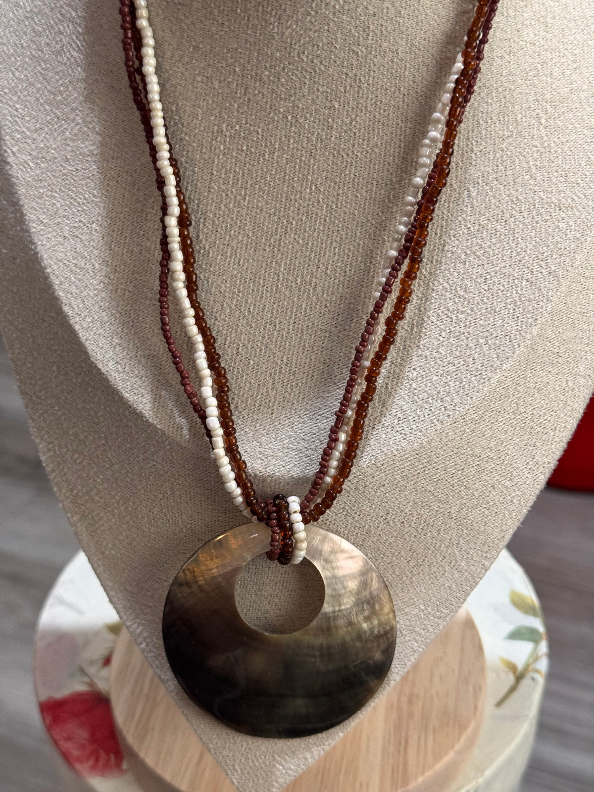 A bohemian-style necklace featuring a large circular shell pendant in a natural brown and black gradient, suspended from two beaded strands in dark brown and cream white, displayed on a beige fabric jewelry bust.