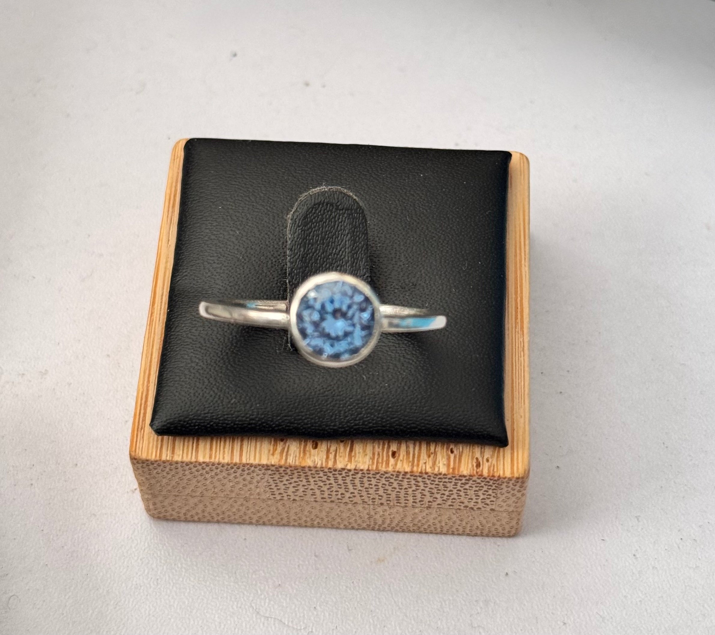 A silver ring with a round blue gemstone set in a bezel setting, displayed on a black cushion inside a bamboo box.