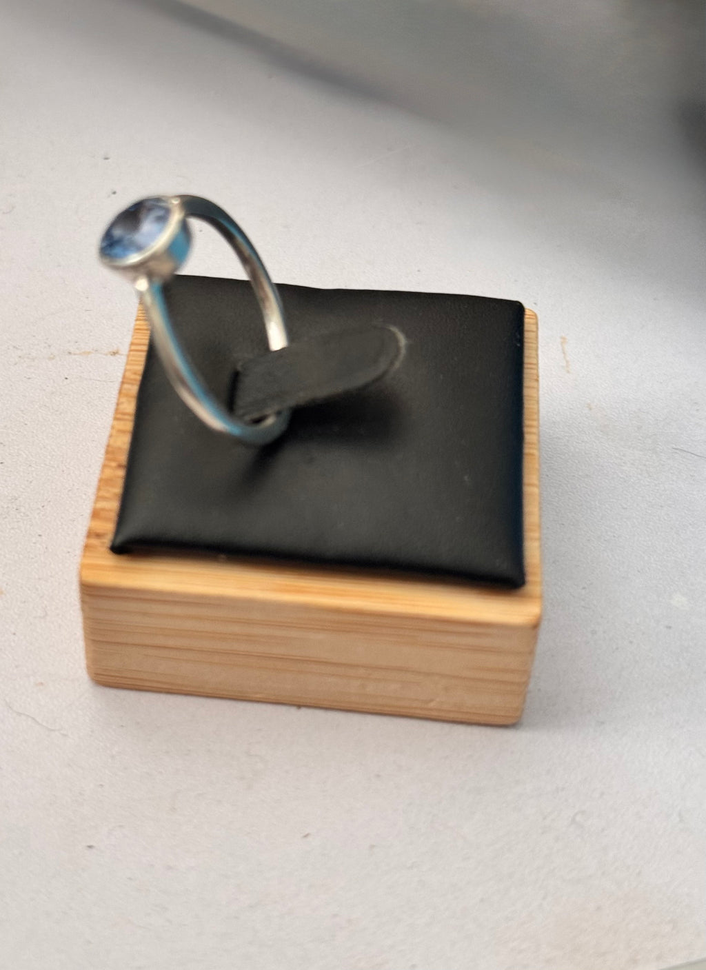 A silver ring with a round blue gemstone displayed on a black cushion atop a light wood block stand against a light gray background. The Left side view