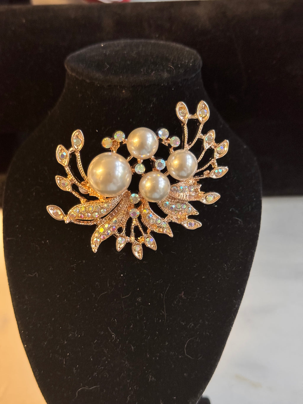 A gold-toned brooch featuring faux pearls and rhinestone-embellished leaves, displayed on a black velvet jewelry bust.